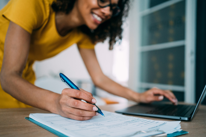 Businesswoman working in the office. Shot of an adorable african-american businesswoman using laptop and making notes on a clipboard inside of the office. Focus is on the pen.